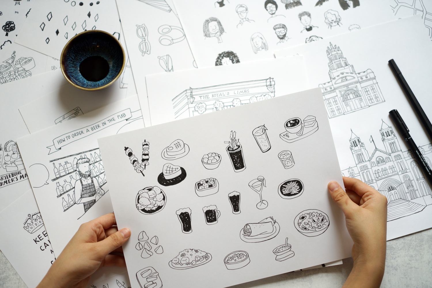 A desk covered in ink drawings, holding up a finished page of food doodles